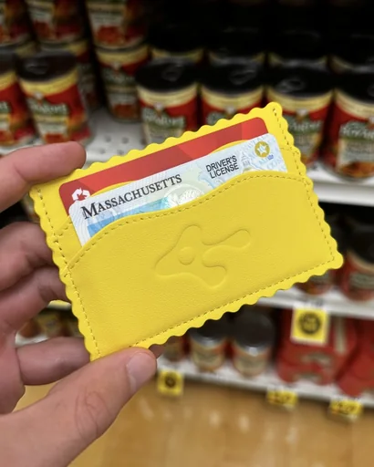 Yellow ravioli-shaped RAVISAUCE card holder held up in a grocery aisle of canned ravioli